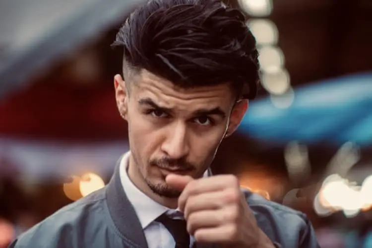 A well dressed man smirking with attitude whilst wearing a tie and white shirt as well as a grey bomber jacket, part of his efforts to keep routines involving self care.
