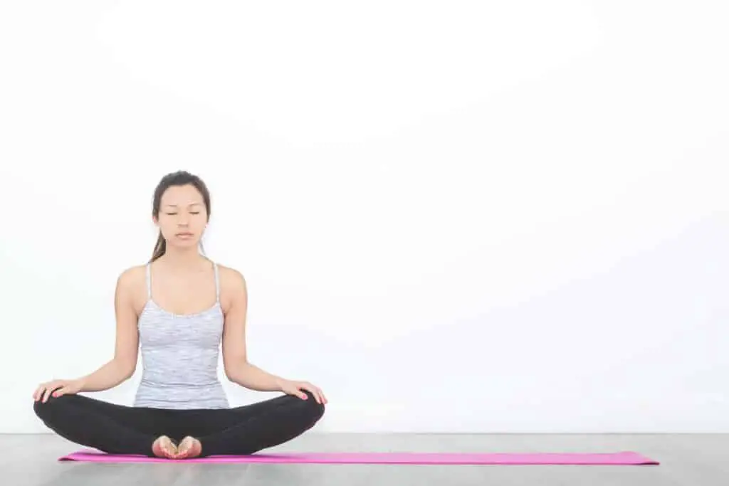 Yoga routines done by a woman sitting and meditating on a pink yoga mat in front of a white background.