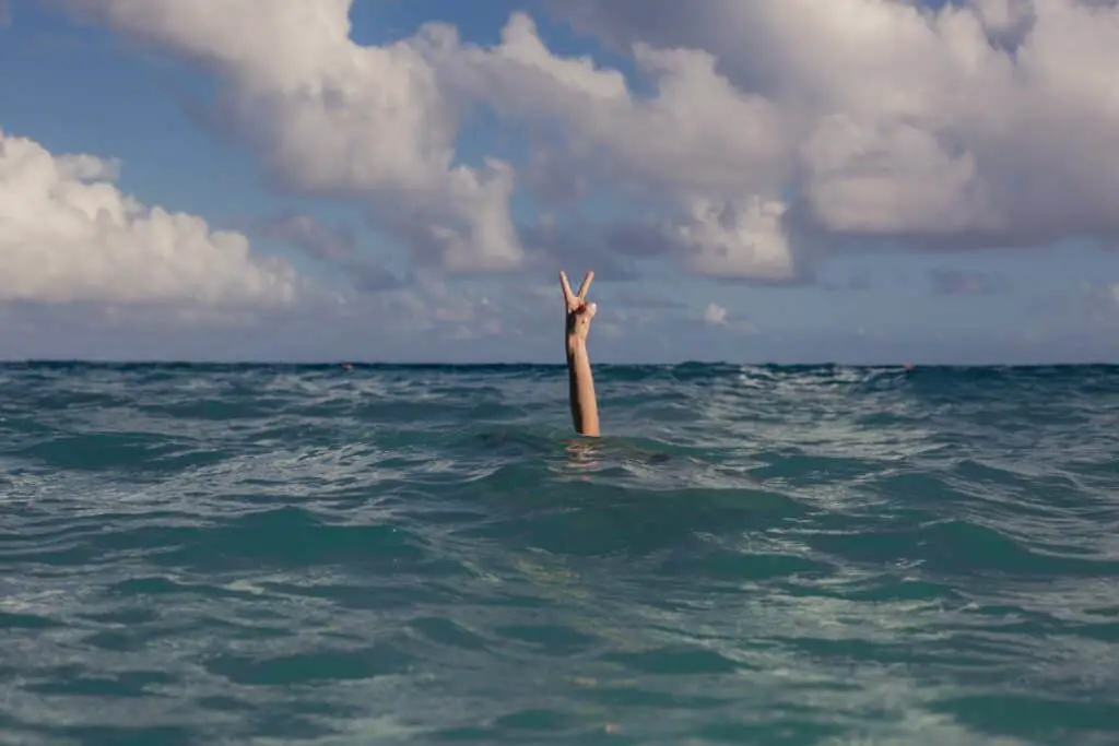 A photo of an arm emerging up from under an empty ocean holding a peace sign just above the waves amongst a horizon of blue nothingness. 