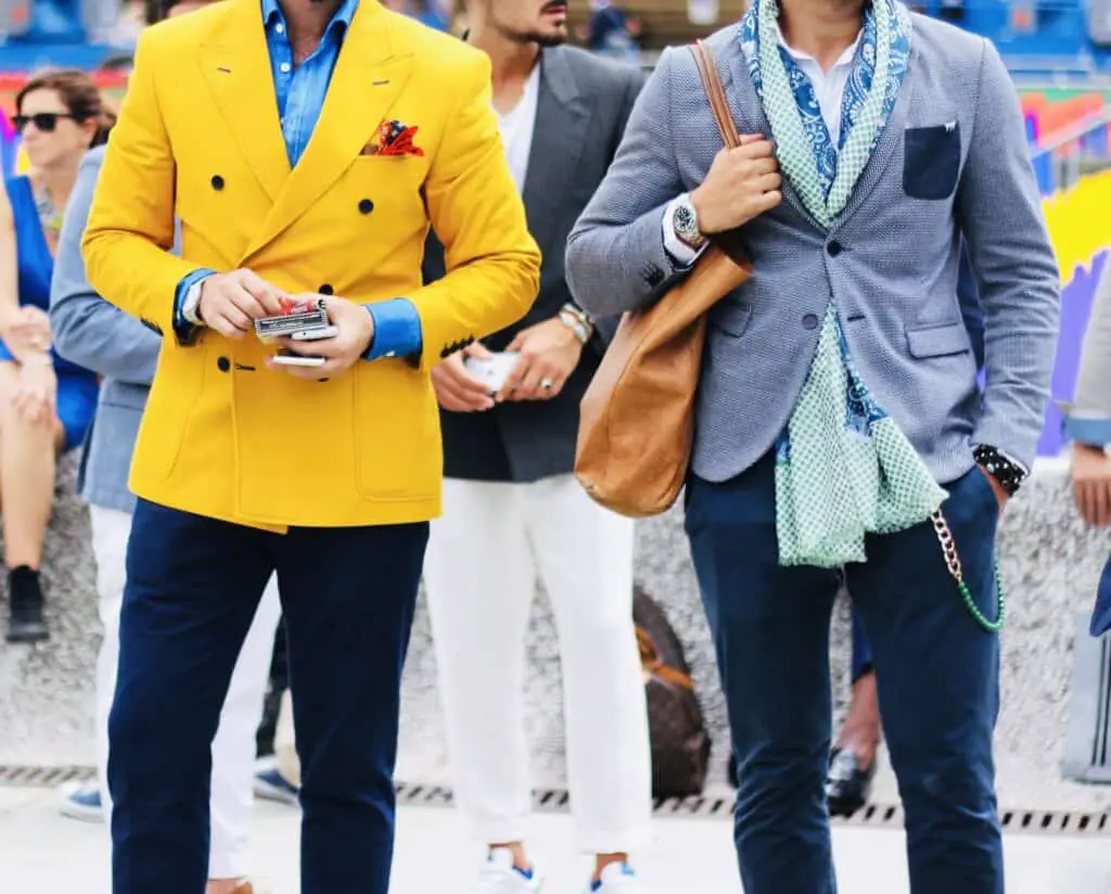 Two stylish men in sartorial clothing, one wearing monochromatic colors while the other wears a bright yellow jacket.
